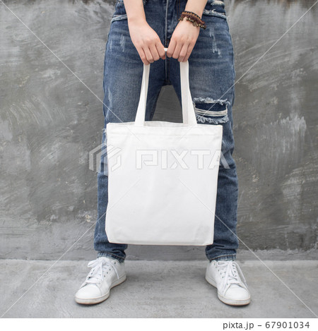 Blank white tote bag canvas fabric with handle mock up design. Close up of woman holding eco or reusable shopping bag on grunge grey wall. No plastic bag and ecology concept. 67901034