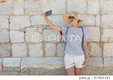 Beautiful young female tourist woman wearing big straw hat, taking self portrait selfie, standing in front of old textured stone wall at old Mediterranean town Beautiful young female tourist woman wearing big straw hat, taking self portrait selfie, standing in front of old textured stone wall at old Mediterranean town 67903193