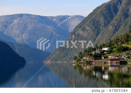 ノルウェー　ハダンゲルフィヨルド、ウルヴィク地区、早朝の朝日に輝く家々と山々と水面の風景を望むアップ 67905584