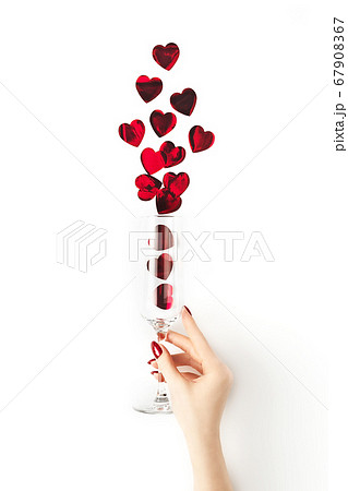 Beautiful female hand with trendy red manicure holding champagne glass with vibrant red heart-shaped confetti poured out of it on white background. Mockup for your design. Copy space. Flat lay. Beautiful female hand with trendy red manicure holding champagne glass with vibrant red heart-shaped confetti poured out of it on white background. Mockup for your design. Copy space. Flat lay. 67908367