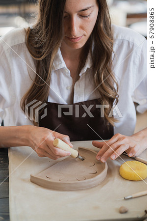 Craftsman hands connect the clay parts made of raw clay, master fingers work with pieces of clay, female sits at a workshop behind the table 67908865