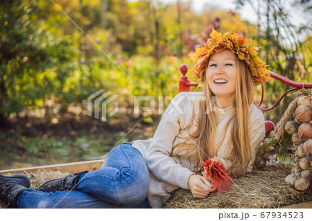 Outdoors lifestyle close up portrait of charming blonde young woman wearing a wreath of autumn leaves on a haystack. Wearing stylish knitted pullover. Wreath of maple leaves Outdoors lifestyle close up portrait of charming blonde young woman wearing a wreath of autumn leaves on a haystack. Wearing stylish knitted pullover. Wreath of maple leaves 67934523