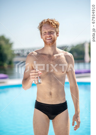 Muscular man with bottle of water standing near pool Muscular man with bottle of water standing near pool 67934910