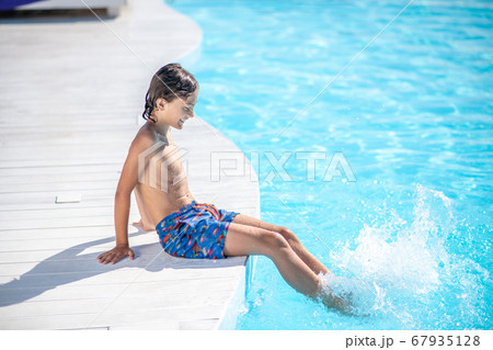 Joyful boy in shorts sitting on poolside of pool 67935128