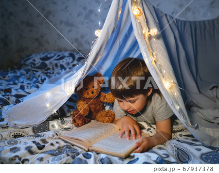 Little boy reads book. Toddler plays in tent made of linen sheet on bed. Cozy evening with favorite book. Little boy reads book. Toddler plays in tent made of linen sheet on bed. Cozy evening with favorite book. 67937378