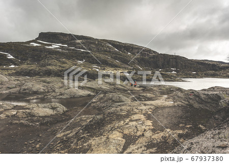 Tourists with backpacks walk across a rocky plain to the famous landmark - Troll tongue rock or Trolltunga. Cold and windy summer in Norway. 67937380