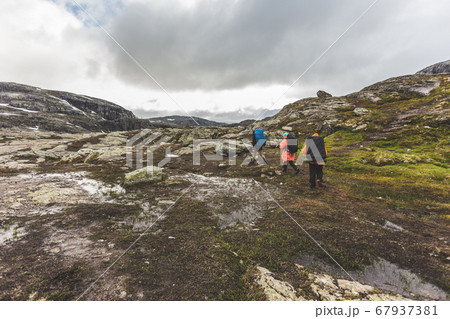 Tourists with backpacks walk across a rocky plain to the famous landmark - Troll tongue rock or Trolltunga. Cold and windy summer in Norway. Tourists with backpacks walk across a rocky plain to the famous landmark - Troll tongue rock or Trolltunga. Cold and windy summer in Norway. 67937381