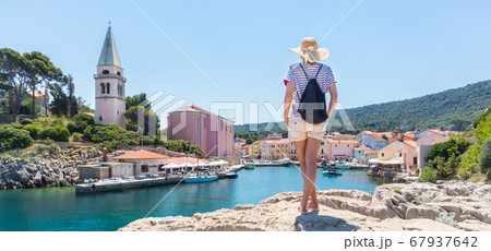 Woman traveler wearing straw summer hat and backpack, standing at edge of the rocky cliff enjoying beautiful panoramic view of Veli Losinj, Losinj island, Croatia 67937642
