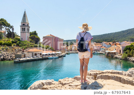 Woman traveler wearing straw summer hat and backpack, standing at edge of the rocky cliff enjoying beautiful panoramic view of Veli Losinj, Losinj island, Croatia 67937643