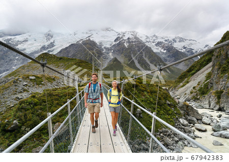 Tourists walking crossing bridge on the Hooker Valley Track hiking trail, New Zealand. Hikers people at Aoraki, Mt Cook National Park with snow capped mountains landscape 67942383