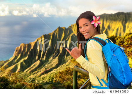 Hawaii hiker girl tourist doing shaka hawaiian hand sign at Na pali lookout in Kauai, Hawaii. Travel Asian woman backpacker hiking with bag at famous destination Kalalau hawaiian attraction Hawaii hiker girl tourist doing shaka hawaiian hand sign at Na pali lookout in Kauai, Hawaii. Travel Asian woman backpacker hiking with bag at famous destination Kalalau hawaiian attraction 67943599