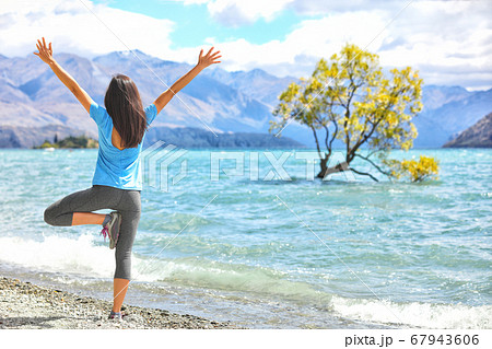 New Zealand morning yoga girl doing tree pose variation doing yoga practice with open arms at Wanaka lake by the lone tree, tourist travel popular attraction in New Zealand. Beach nature landscape 67943606