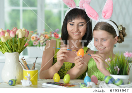 Happy mother with daughter wearing rabbit ears decorating Easter eggs 67953947