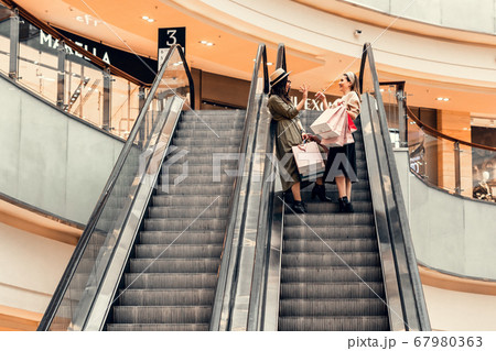 Two girls on an escalator in a shopping center. In the hands of paper bags. Shopping, leisure and entertainment. 67980363
