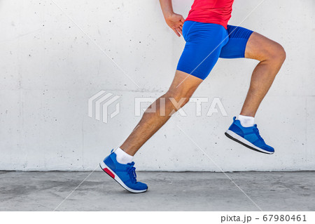 Fitness athlete man running on asphalt sidewalk in city street. Sport lifestyle. Closeup of lower body runner's legs on workout marathon run against white wall background. Shoes, compression clothing. 67980461