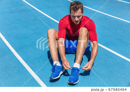 Athlete sprinter getting ready to run tying up shoe laces on stadium running tracks. Man runner preparing for cardio training outdoors. Fitness and sports. 67980464