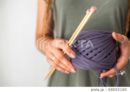A young girl knitter with dreadlocks in a green dress holds a skein of violet yarn and wooden knitting needles for knitting. Light background. A young girl knitter with dreadlocks in a green dress holds a skein of violet yarn and wooden knitting needles for knitting. Light background. 68003160