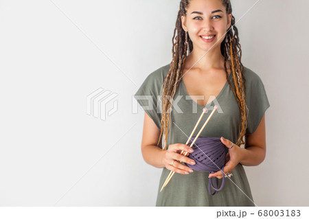 A young girl knitter with dreadlocks in a green dress holds a skein of blue yarn and wooden knitting needles for knitting. Light background. 68003183