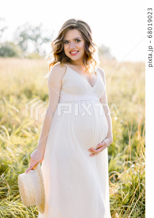 Attractive young pregnant european woman in white dress, touching her big beautiful belly and walking in the meadow with sunset 68019023