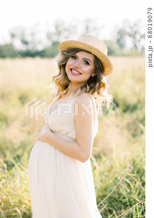 Smiling charming young pregnant woman, wearing white dress and straw hat, resting in summer field. Posing outdoors. Motherhood. Outdoor portrait of beautiful pregnant woman in white dress 68019079