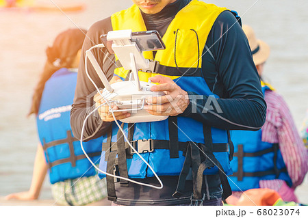 Young man in blue and yellow life vest controlling a drone to take pictures of ocean coast with tourists while traveling on a raft in the sea. Hands holding drone remote controller. 68023074