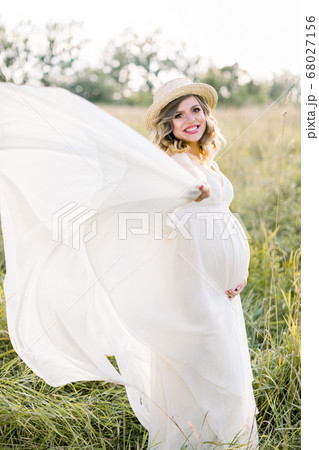 Smiling pregnant woman in white dress and straw hat, walking and having fun in summer field. Pregnant woman outdoors in countryside. Happy woman in field outdoors 68027156