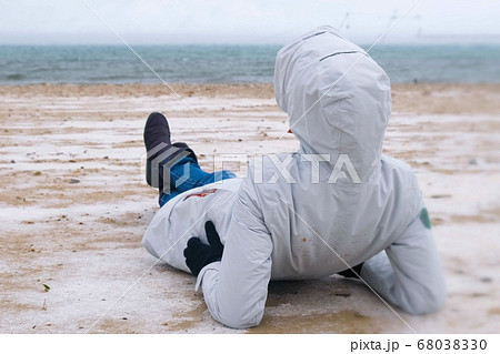 Woman tourist lies on a snowy sandy beach in winter by the sea. Woman in a jacket and jeans, back view. 68038330