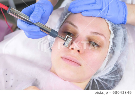 Cosmetologist makes a micro-current procedure for the face of a young woman. Face close-up, front view. 68043349