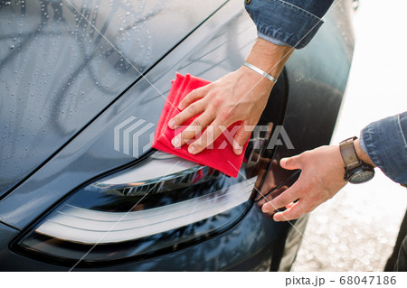 Closeup cropped image of hands of young man, washing and cleaning his modern blue car with red microfiber cloth, wiping hood and headlights. Car wash outdoors concept 68047186