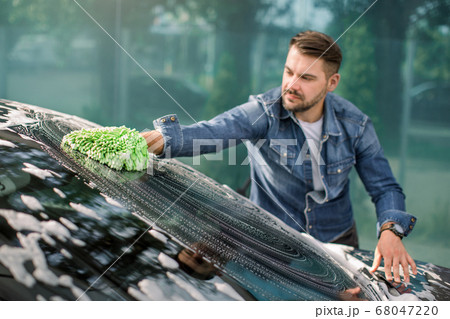 Horizontal view of car cleaning at self wash service outdoors. Handsome Caucasian man in casual wear washing the car windshield with green mitten sponge and foam. 68047220