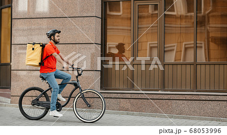 Delivery, work and sport. Young guy with beard in helmet with backpack rides a bicycle down the street 68053996