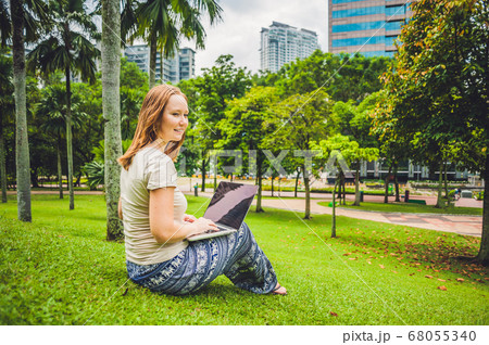 A young woman in casual dress using laptop in a tropical park on the background of skyscrapers. Mobile Office concept 68055340