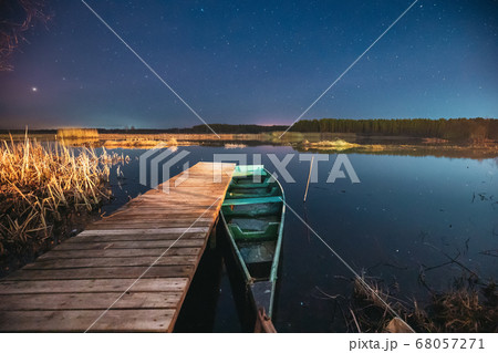 Belarus, Eastern Europe. Real Night Sky Stars Above Old Pier With Moored Wooden Fishing Boat. Natural Starry Sky And Countryside Landscape With Lake River In Early Spring Night. Russian Nature 68057271