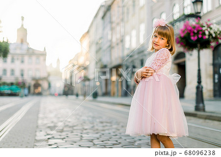 Horizontal shot of little Caucasian blond girl in a beautiful dress standing in ancient city center outdoors. City lifestyle portrait, fashionable concept. Copy space 68096238