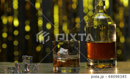 Golden whiskey, cognac or brandy in glass with ice cubes and bottle on table. Shiny background Golden whiskey, cognac or brandy in glass with ice cubes and bottle on table. Shiny background 68096385