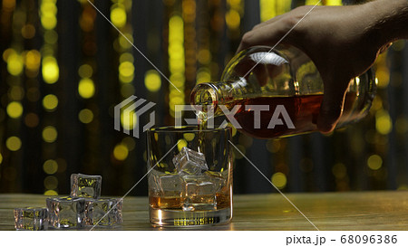 Pouring of golden whiskey, cognac or brandy from bottle into glass with ice cubes. Shiny background Pouring of golden whiskey, cognac or brandy from bottle into glass with ice cubes. Shiny background 68096386