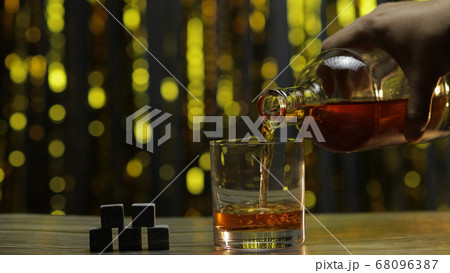 Pouring of golden whiskey, cognac or brandy from bottle into glass with rocks ice stones on table Pouring of golden whiskey, cognac or brandy from bottle into glass with rocks ice stones on table 68096387