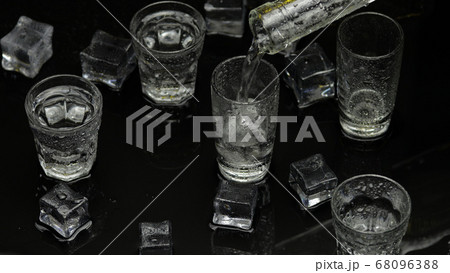 Barman pour frozen vodka from bottle into shot glass. Ice cubes against dark wet black background Barman pour frozen vodka from bottle into shot glass. Ice cubes against dark wet black background 68096388