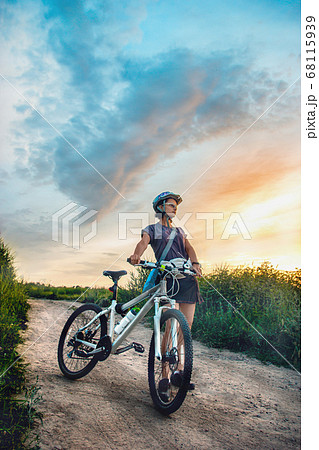 Young woman and mountain bike stands in the rural green field at summer evening. Adventure summer leisure time. 68115939
