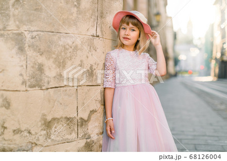 Cute little girl in fashionable pink hat and dress posing to camera outdoors in the city street, standing near old stone wall of ancient building in European city Cute little girl in fashionable pink hat and dress posing to camera outdoors in the city street, standing near old stone wall of ancient building in European city 68126004