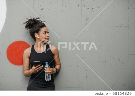 Muscular african american girl with modern gadgets for workout holds bottle and smartphone on gray wall background in city 68154829