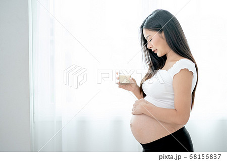 Beautiful Asian pregnant woman hold glass of milk and look to her belly in front white curtain 68156837