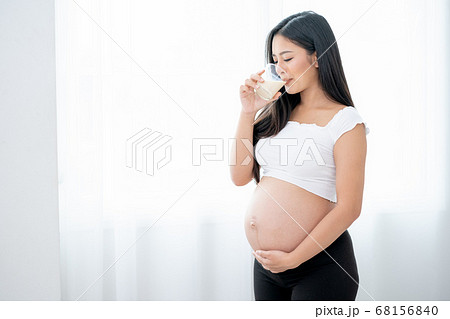 Beautiful Asian pregnant woman drink milk and stand in front of white curtain and she look happy Beautiful Asian pregnant woman drink milk and stand in front of white curtain and she look happy 68156840