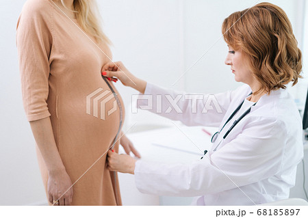 Serious female doctor wearing white coat measure belly waist size of pregnant young woman Serious female doctor wearing white coat measure belly waist size of pregnant young woman 68185897