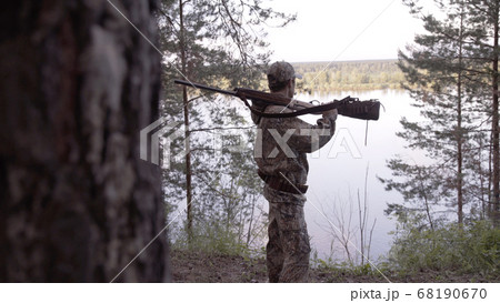 View from back of hunter in camouflage puts hunting rifle on his shoulder and looks at river from forest. Outdoor activity concept. Summer hiking in wilderness area. View from back of hunter in camouflage puts hunting rifle on his shoulder and looks at river from forest. Outdoor activity concept. Summer hiking in wilderness area. 68190670