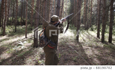 Man takes aim while standing from hunting rifle. Man in comfortable camouflage clothes hunter outdoor in forest hunting alone. Hunter in camouflage aims gun at object in forest during sunset. 68190702