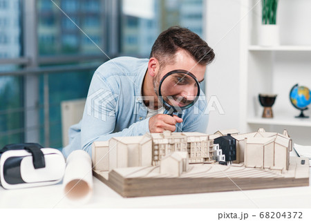 Funny picture of male architect looks through magnifying glass to a house model. House inspection and real estate concept. 68204372