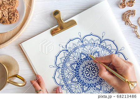 Female hands close up draw Decorative round floral mandala. Hobby and home relaxation. A mug of coffee and cookies on a wooden tray. White background. Female hands close up draw Decorative round floral mandala. Hobby and home relaxation. A mug of coffee and cookies on a wooden tray. White background. 68213486