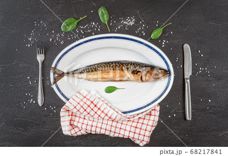 Whole smoked mackerel fish on white oval plate, gray black stone like table under, rock salt and green leaves salad near. View from above Whole smoked mackerel fish on white oval plate, gray black stone like table under, rock salt and green leaves salad near. View from above 68217841