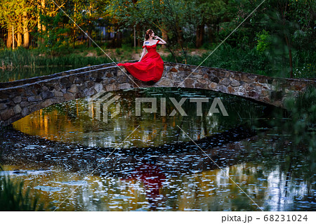 Redhead woman in red dress sitting on stone bridge 68231024